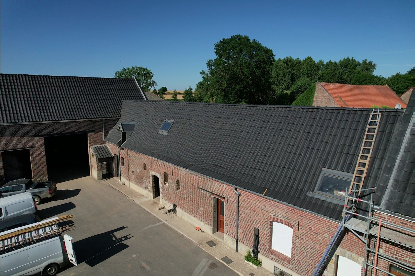 Bâtiment en briques avec toit en tuiles noires, cour, camionnettes garées, arbres et ciel bleu.