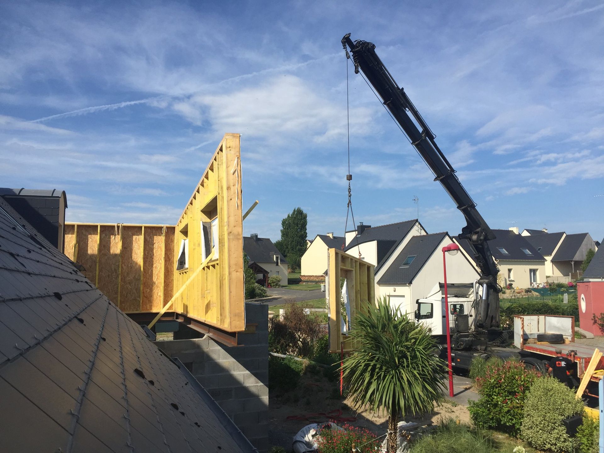 Une grue de chantier soulève un élément de mur préfabriqué en bois.