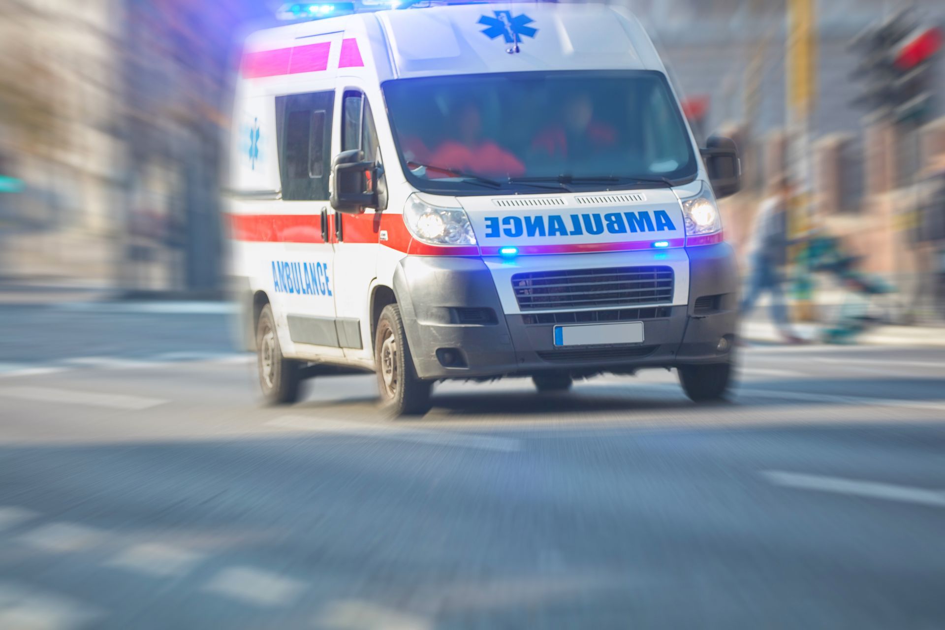 Photo d'une ambulance de face blanche rouge et bleue en train de rouler. Le paysage en arrière-plan est flou pour donner l'impression de vitesse.