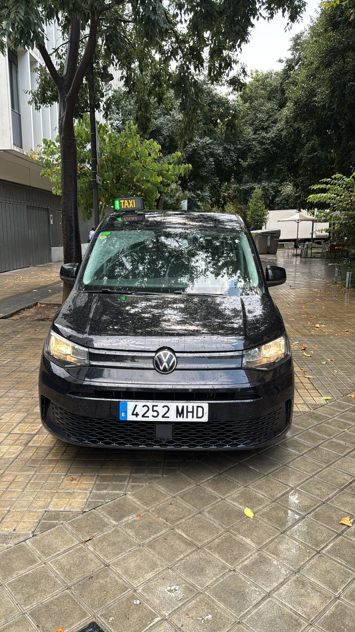 Taxi negro estacionado en una calle adoquinada mojada, matrícula 6237 MHJ.