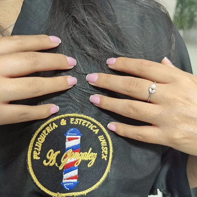 Una mujer con uñas rosas lleva un anillo y el logo de una barbería.