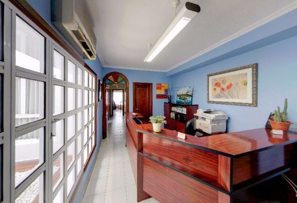 Pasillo de oficina luminoso con ventanas de cristal, mostrador de recepción de madera, paredes azules y luces fluorescentes.