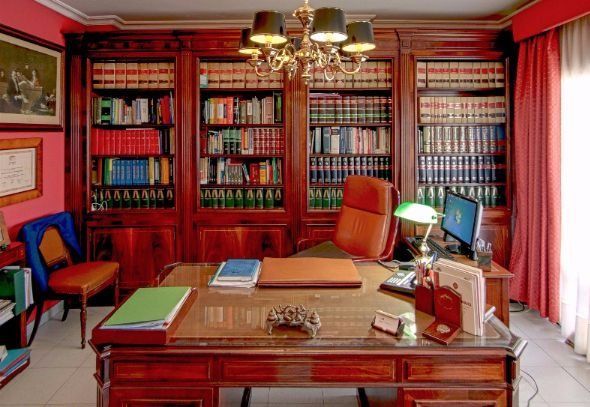 Elegante despacho en casa con estanterías rojas, escritorio de madera y ordenador junto a una ventana soleada.