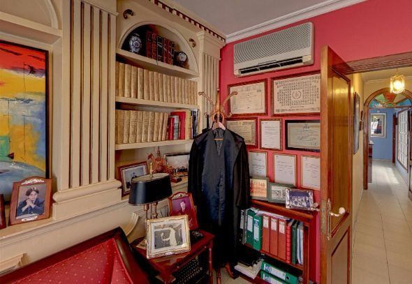 Acogedor pasillo con estanterías, documentos enmarcados, un abrigo colgado y paredes rojas junto a una puerta.