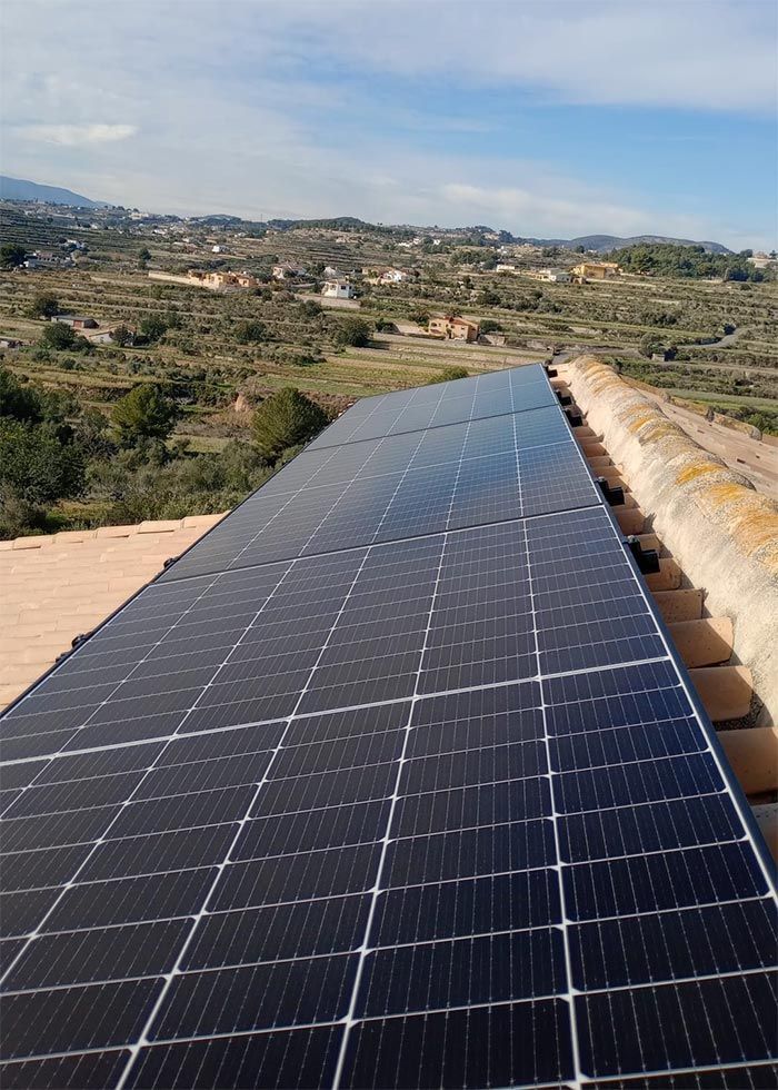 Paneles solares sobre un techo de tejas de terracota con un paisaje rural al fondo bajo un cielo azul.