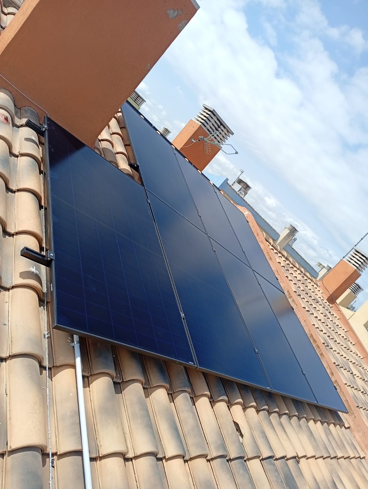 Paneles solares montados sobre un tejado de tejas, chimeneas visibles contra un cielo parcialmente nublado.