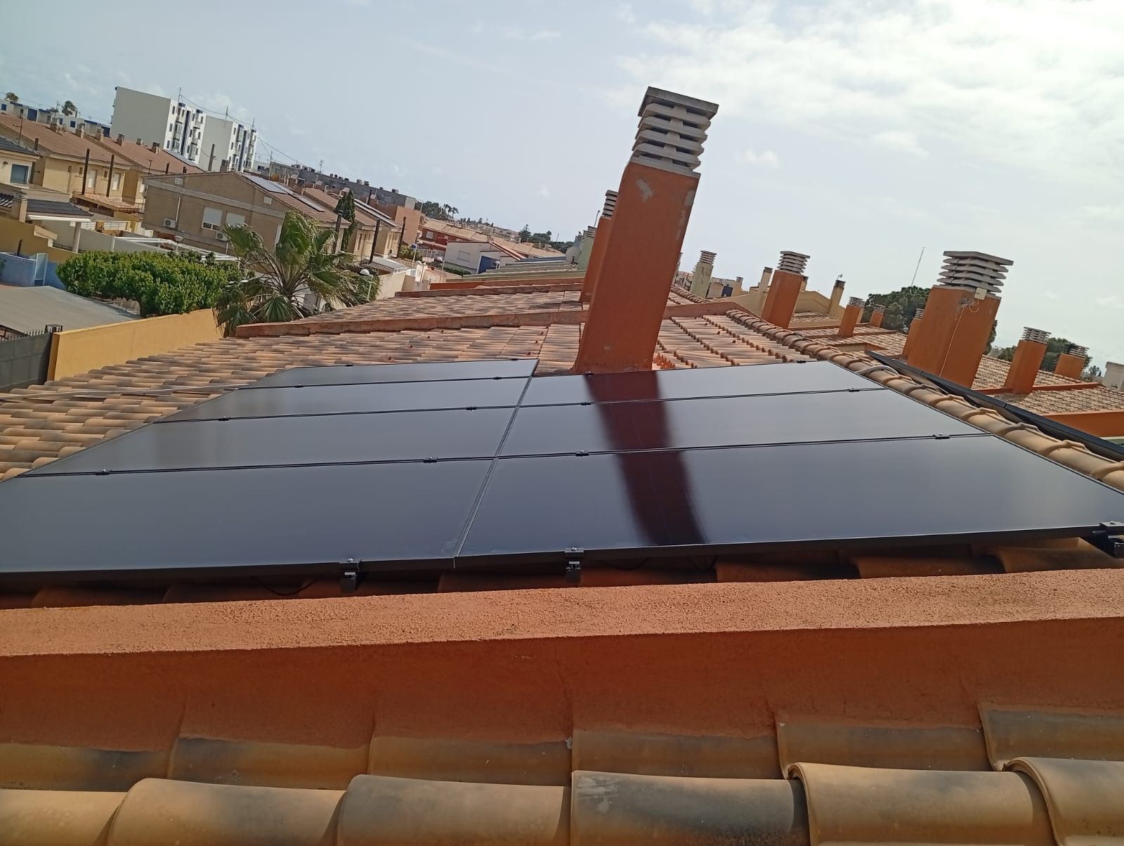 Paneles solares sobre un tejado de tejas de terracota con chimeneas, sobre un fondo de edificios bajo un cielo soleado.
