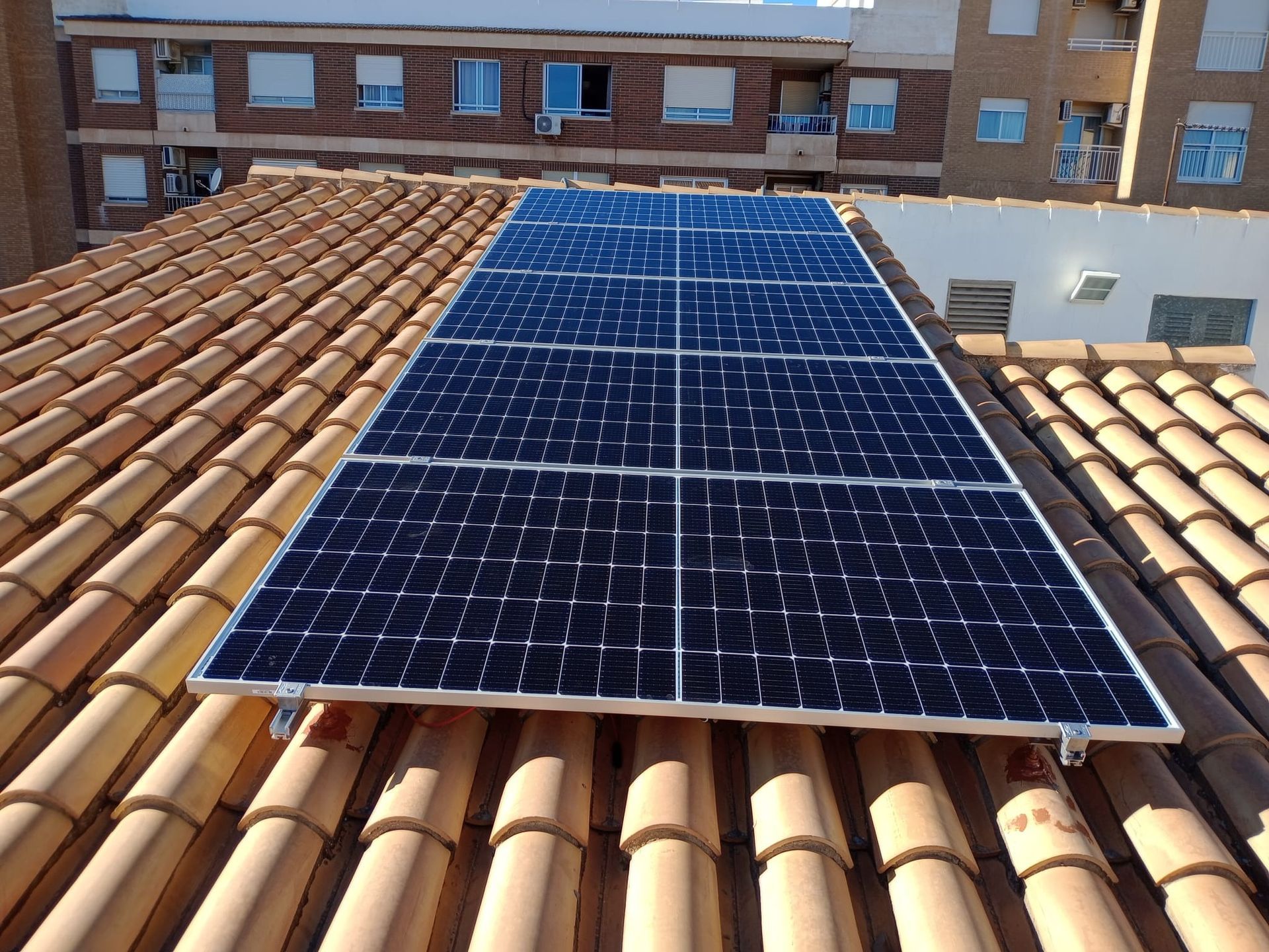 Paneles solares instalados sobre un tejado de tejas de terracota, con un edificio al fondo.