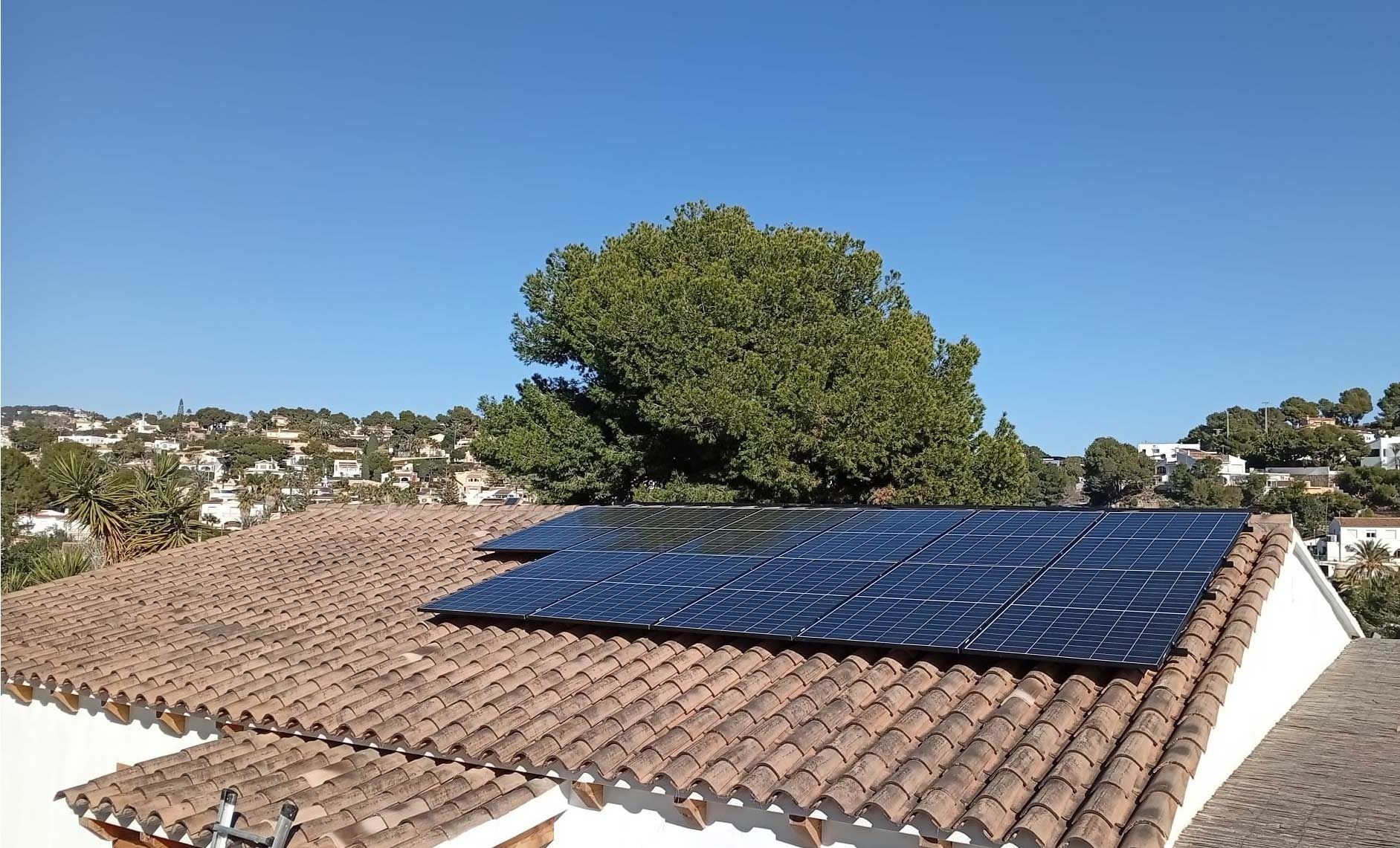 Paneles solares sobre un techo de tejas de terracota con un árbol y casas al fondo bajo un cielo azul.