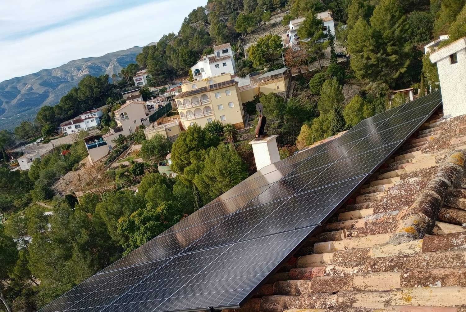 Paneles solares sobre un tejado de tejas de terracota en un pueblo en la ladera de una montaña; árboles y edificios al fondo.