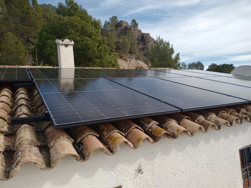 Paneles solares instalados en un tejado de tejas, chimenea, árboles y cielo parcialmente nublado al fondo.