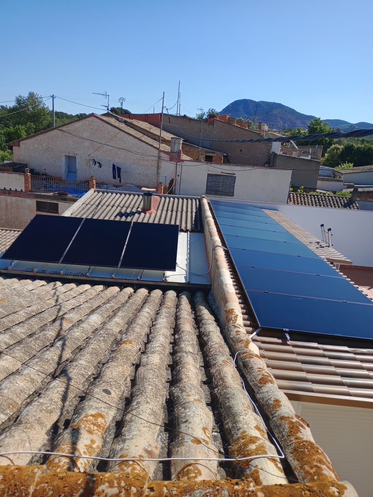 Paneles solares sobre un tejado de tejas. Cielo azul con edificios y montañas al fondo.