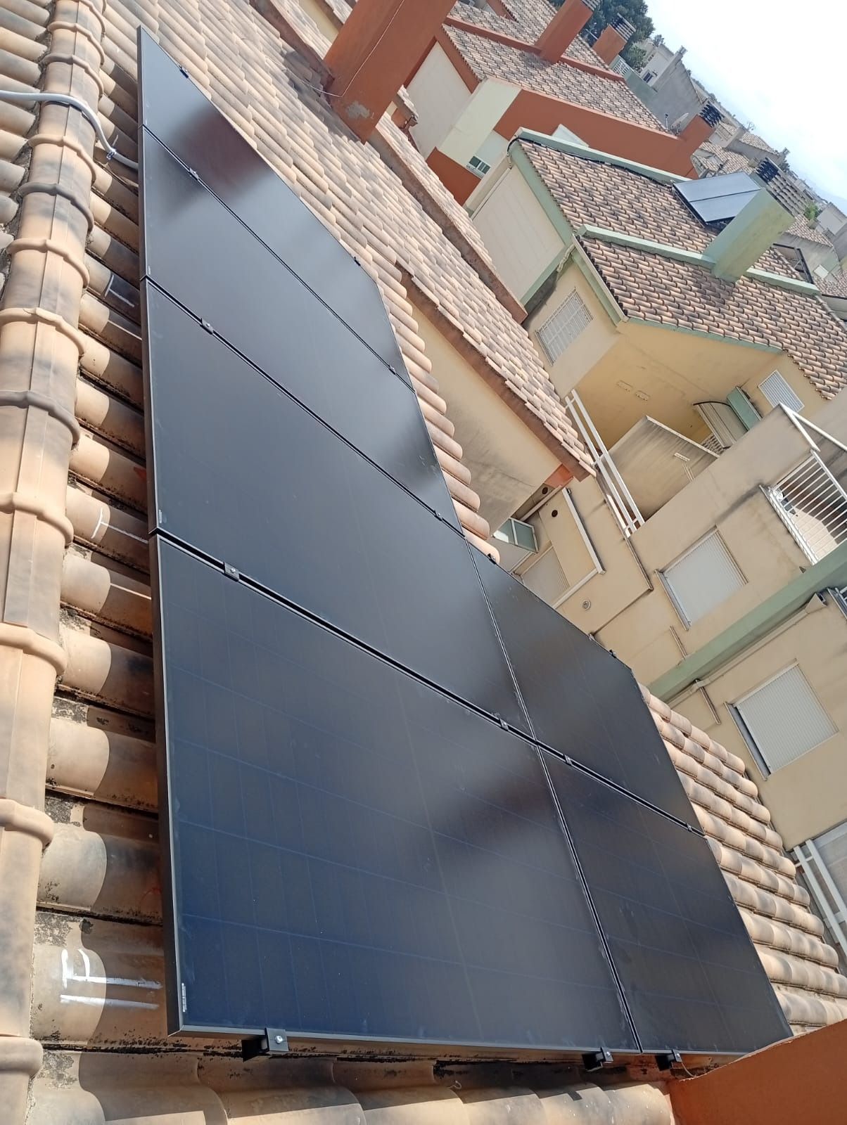 Paneles solares instalados en un tejado de terracota, orientados hacia el sol. Edificio de apartamentos al fondo.