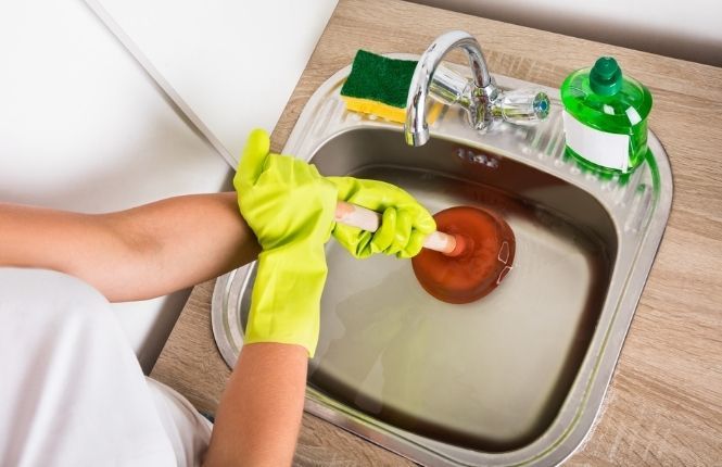 Eine Person benutzt einen Pömpel, um das Wasser aus einem Waschbecken zu entleeren.