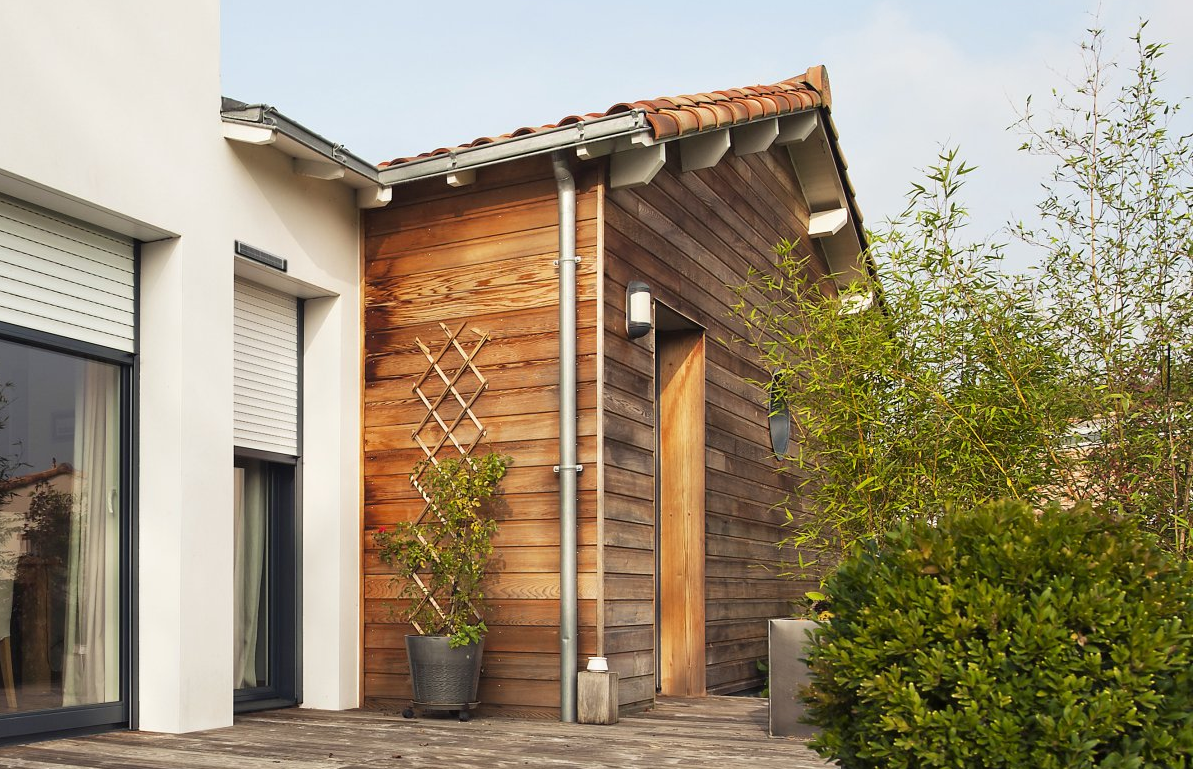 Vue extérieure d'une maison avec une dépendance en bois. On y voit des plantes et une porte fermée.