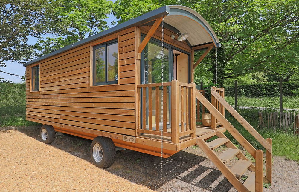 Cabane en bois sur roues avec petit porche, marches et deux fenêtres. Toit gris, bardage brun clair, roues visibles.