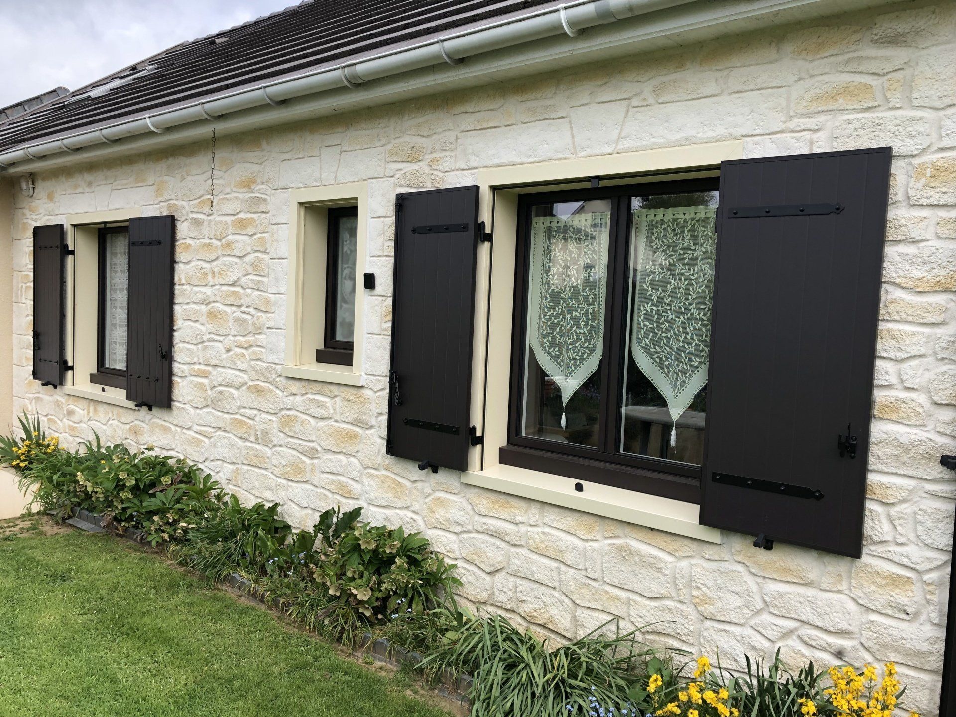 Volets noirs sur une maison en pierre claire, pelouse verte et arbustes devant.