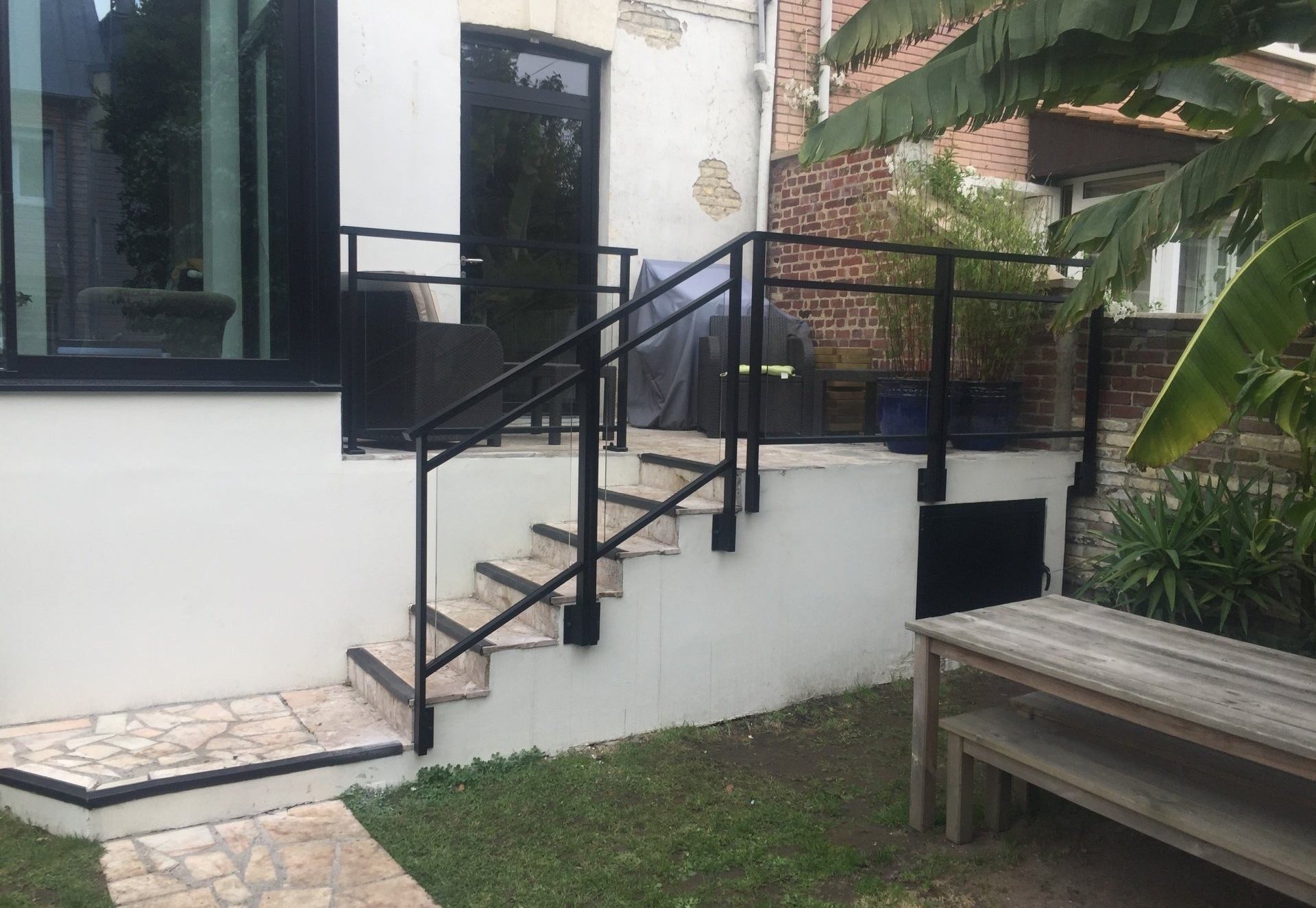 Rambarde en métal noir sur un escalier extérieur menant à une terrasse. Bâtiment blanc, banc en bois, pelouse verte.