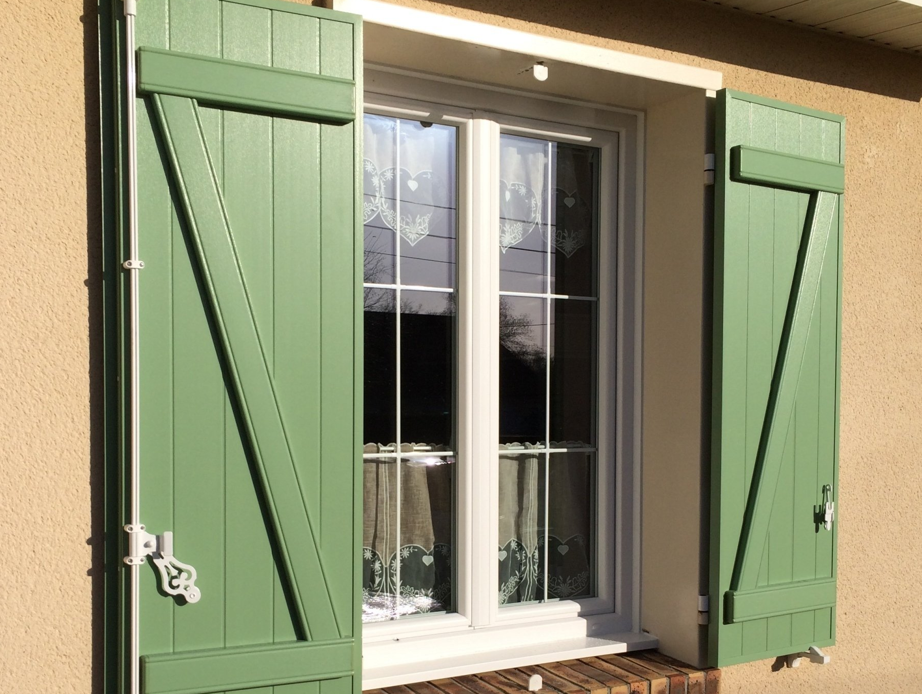 Des volets verts encadrent une fenêtre à cadre blanc sur un mur beige. La lumière du soleil se reflète sur la vitre.