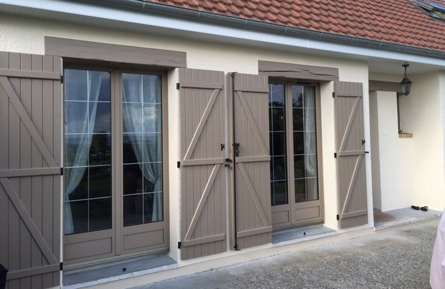 Volets beiges sur un bâtiment couleur crème, deux paires de portes-fenêtres, toit de tuiles, terrasse en pierre.