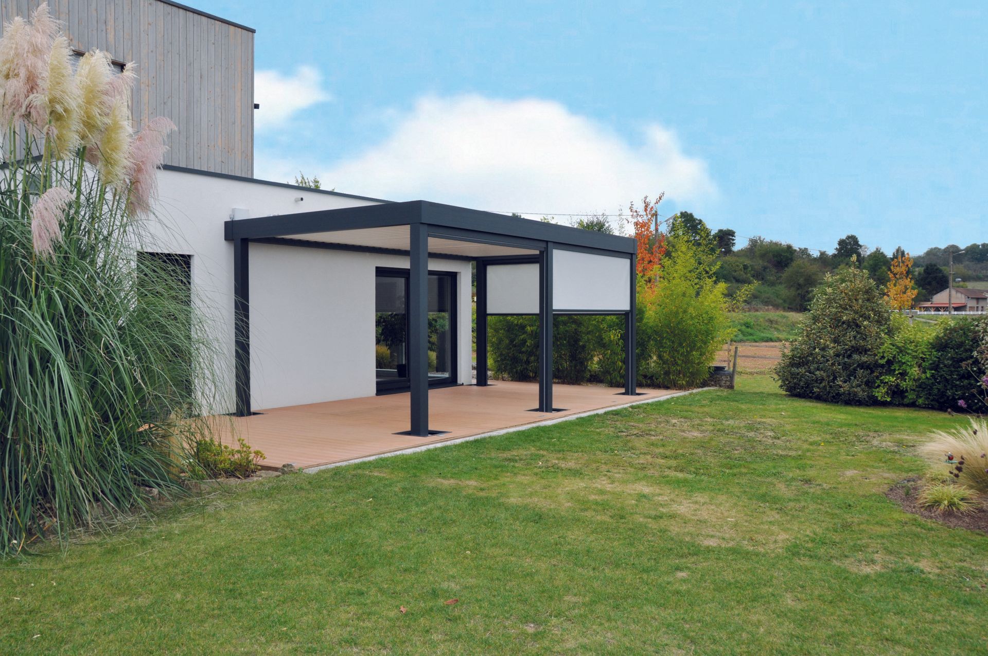Pergola sur terrasse avec murs blancs, colonnes noires et stores enrouleurs. Pelouse verte et arbres tout autour.