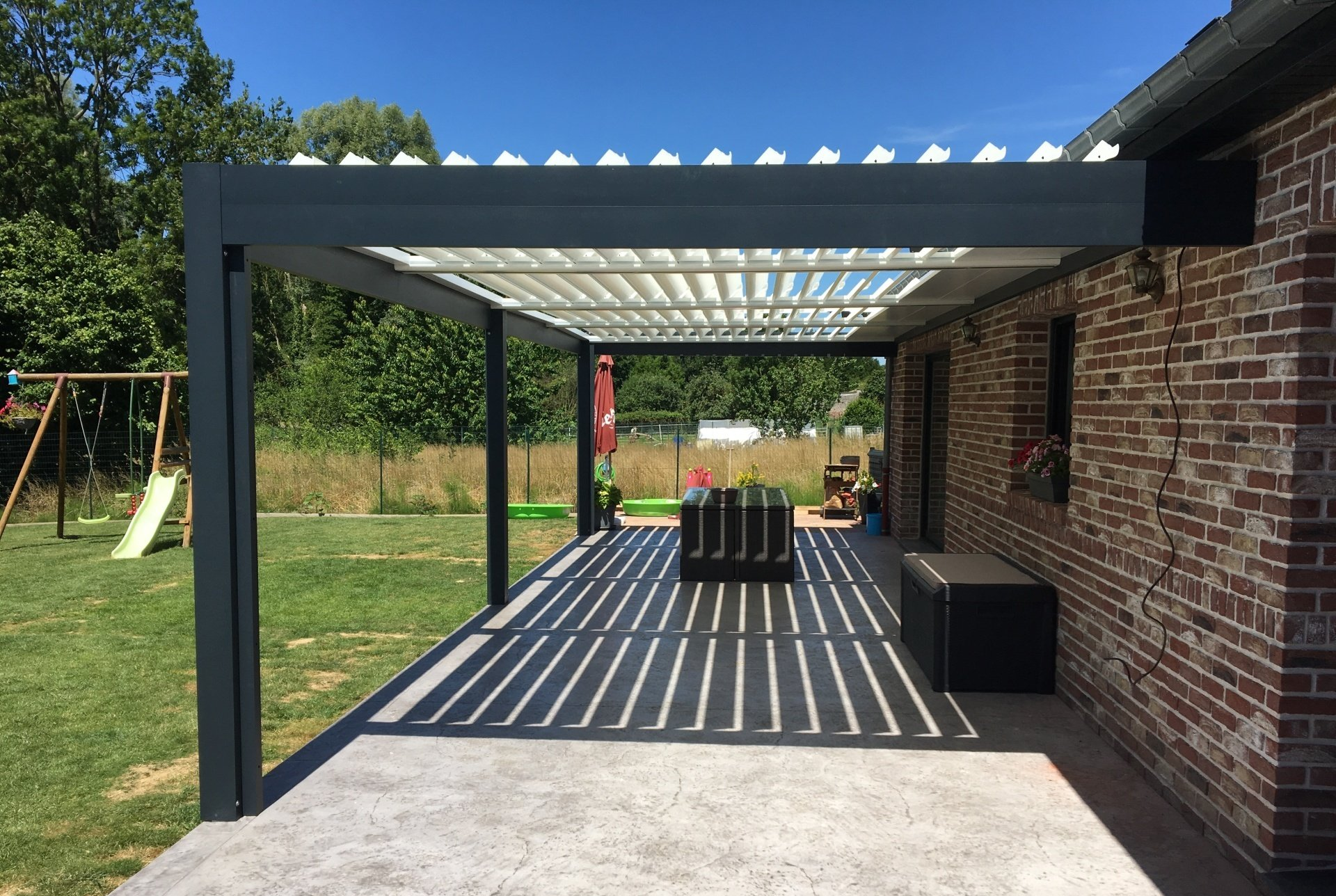 Une pergola surplombe une terrasse en béton jouxtant une maison en briques. De longues ombres se projettent sur la terrasse.
