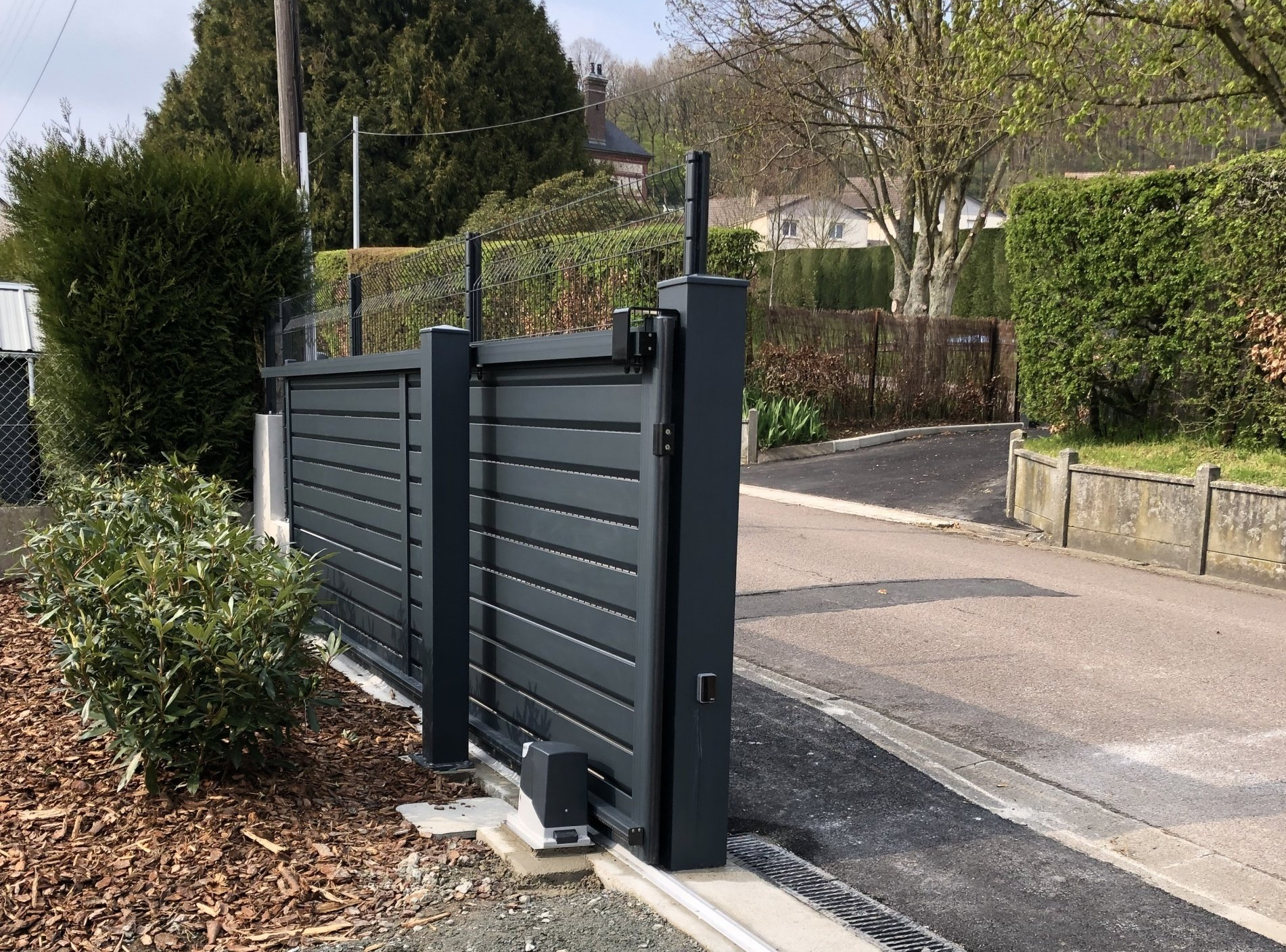 Portail coulissant gris foncé, partiellement ouvert, donnant sur une route et des buissons.