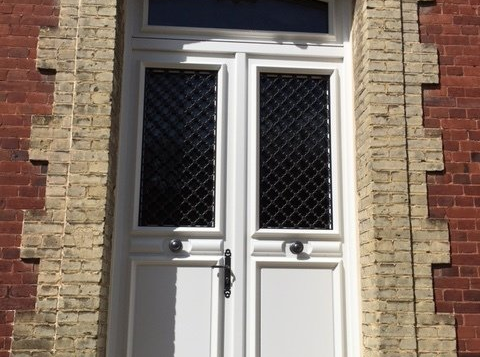 Porte double blanche vitrée, ferronnerie noire, encadrée de briques claires, et bâtiment en briques rouges.