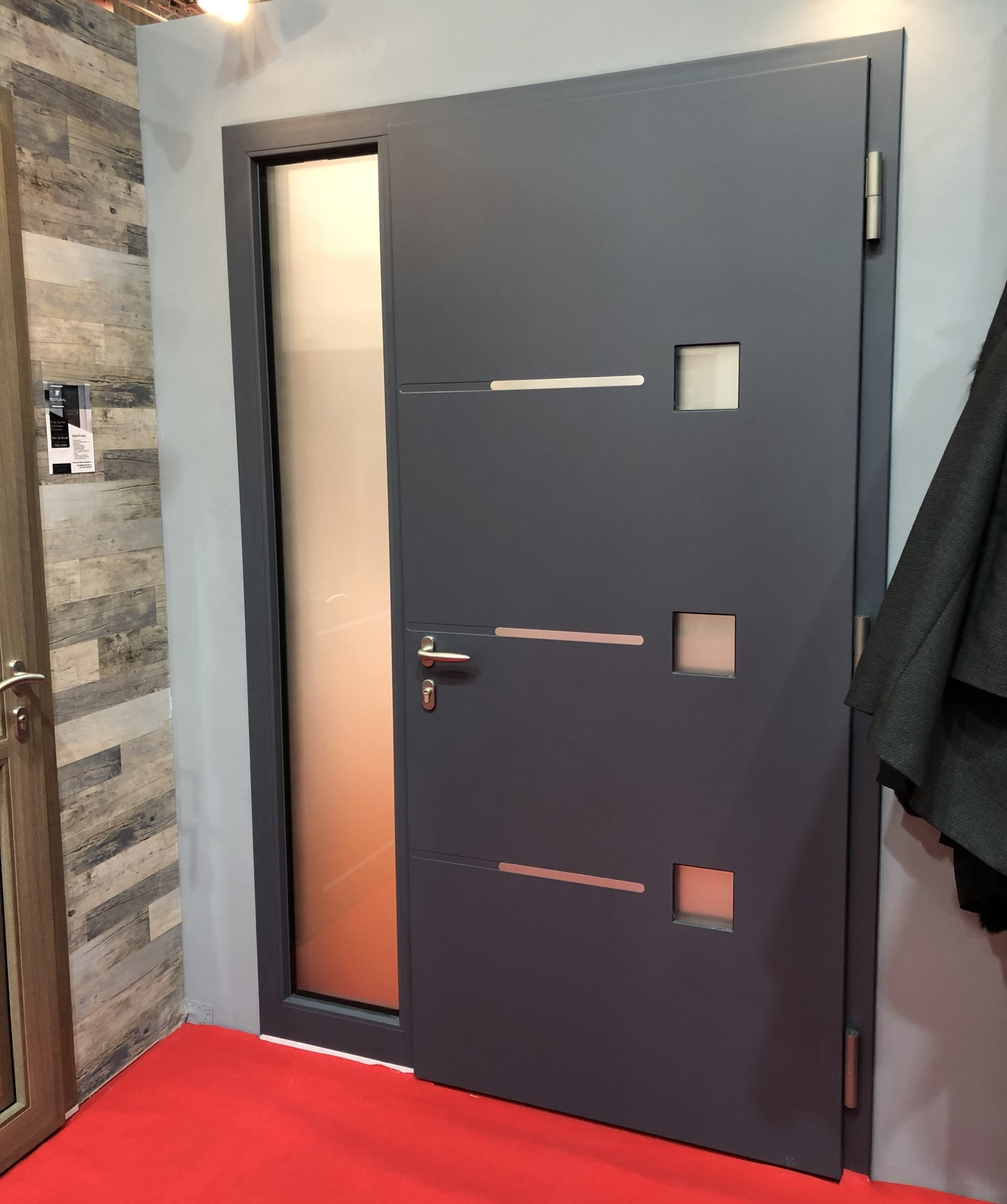 Porte d'entrée moderne grise avec imposte latérale et motifs ajourés. Tapis rouge au sol.