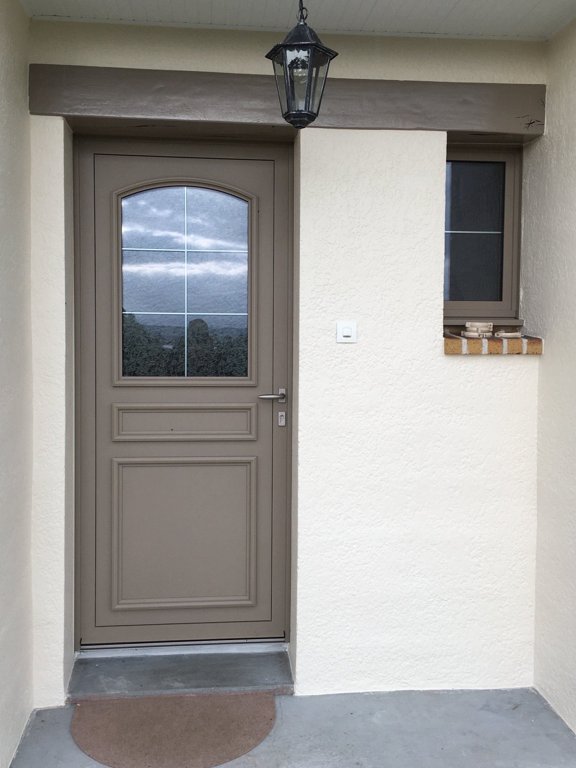 Porte beige avec panneau de verre, encadrement marron, petite fenêtre et éclairage extérieur.