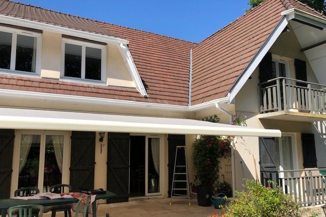 Maison beige avec toit marron et auvent au-dessus d'une terrasse.