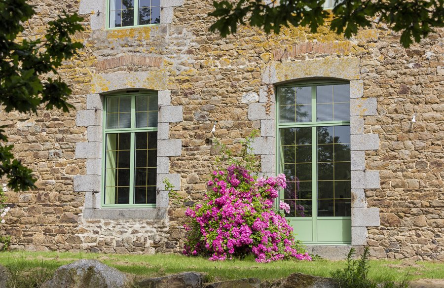 Bâtiment en pierre aux fenêtres à encadrement vert, fleurs roses devant et feuillage au-dessus.