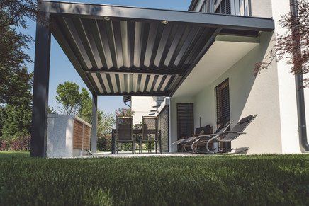 Pergola moderne attenante à une maison blanche avec mobilier de jardin sur une terrasse ; pelouse verte au premier plan.