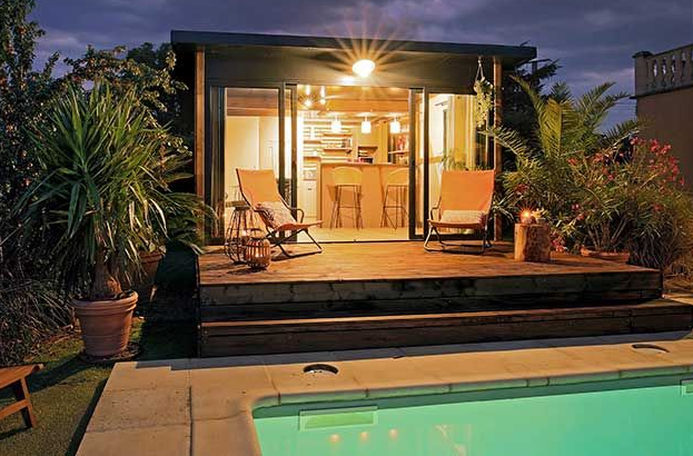 Pergola au-dessus d'une terrasse, projetant des ombres aux motifs géométriques sur la pierre. Adossée à une maison en briques