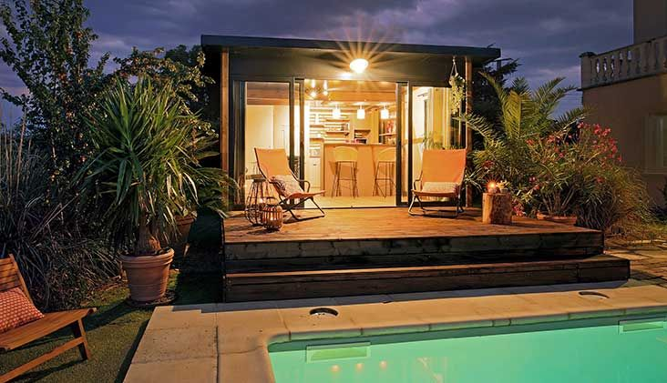 Cabane au bord de la piscine au crépuscule, portes ouvertes, chaises et éclairage intérieur chaleureux.