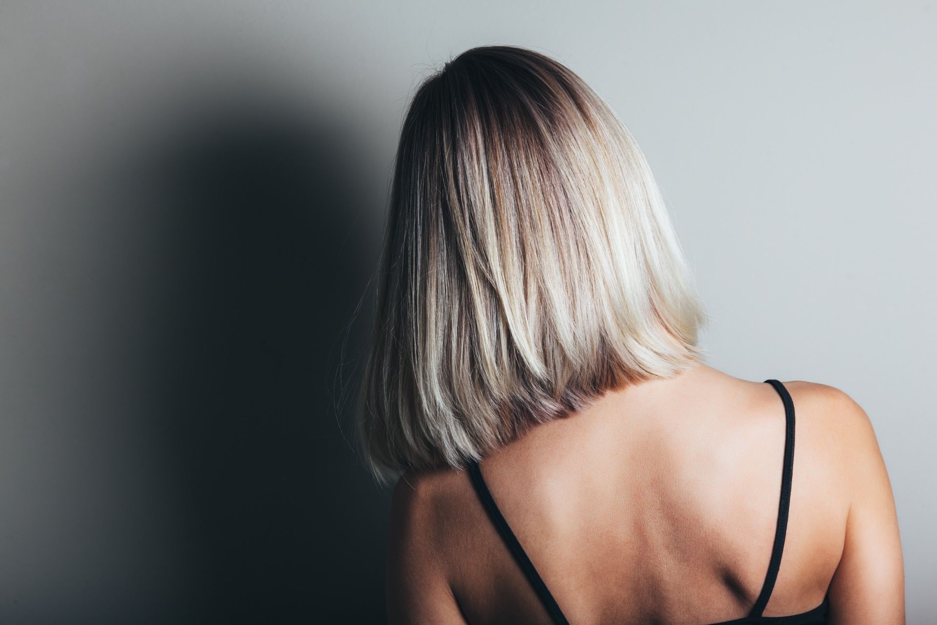 Mujer con cabello rubio y texturizado, de espaldas a la cámara, vistiendo una camiseta negra sin mangas, contra una pared gris.