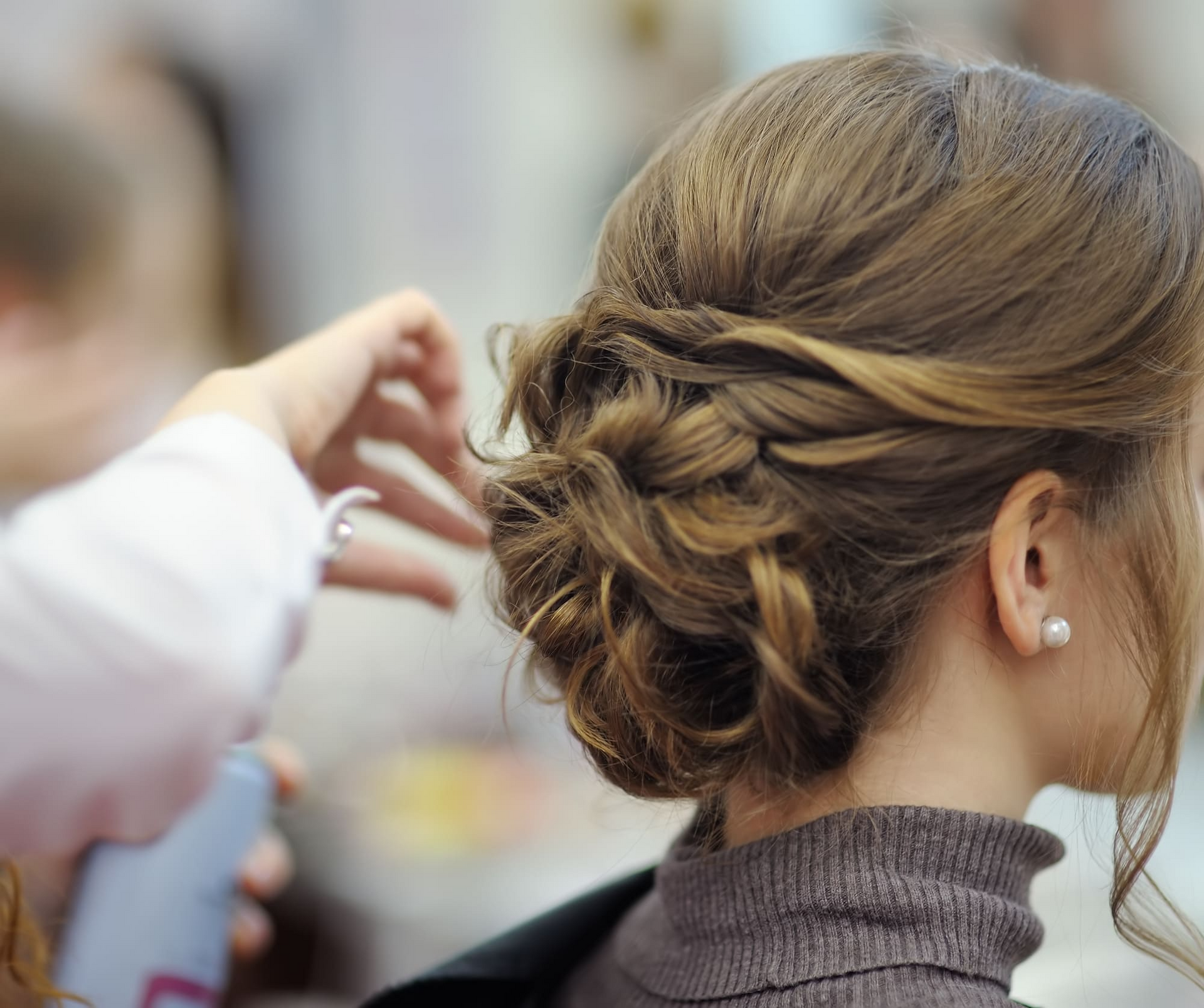 Mujer haciéndose un moño; mano del estilista, cabello castaño, cuello alto gris, pendiente de perla.