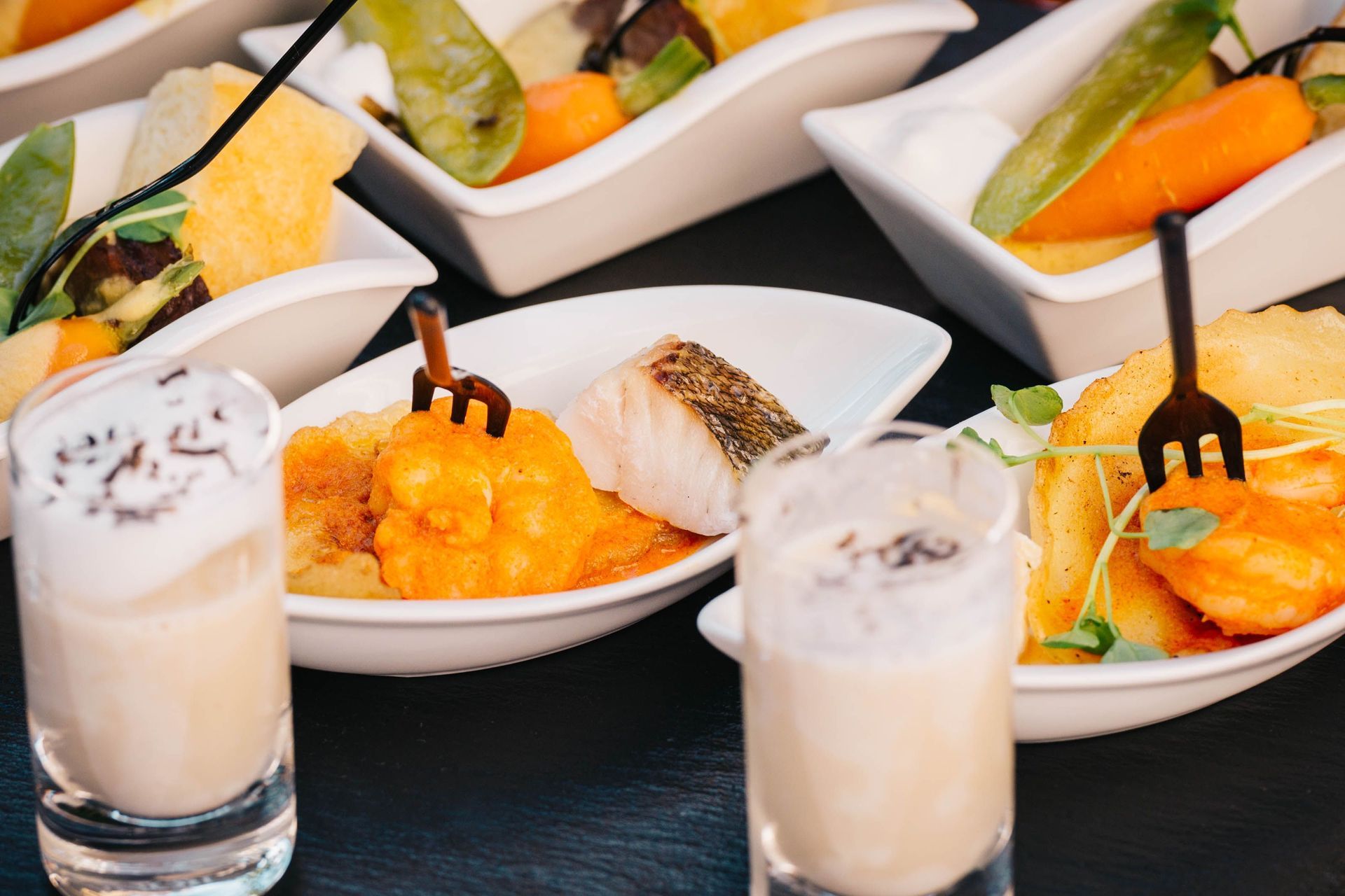 Assortiment de hors-d'œuvre gastronomiques dans des plats blancs, accompagnés de deux boissons crémeuses sur une table sombre.