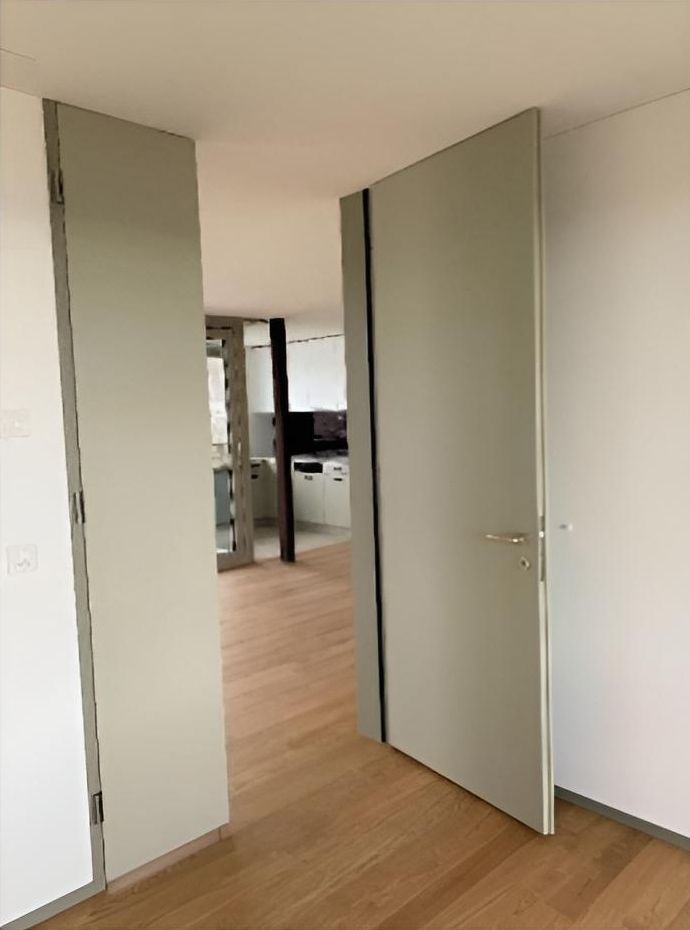 Eine offene, hellgraue Innentür mit einem schmalen, dunklen, vertikalen Türgriff gibt den Blick auf einen Holzboden und den dahinterliegenden Küchenbereich frei.