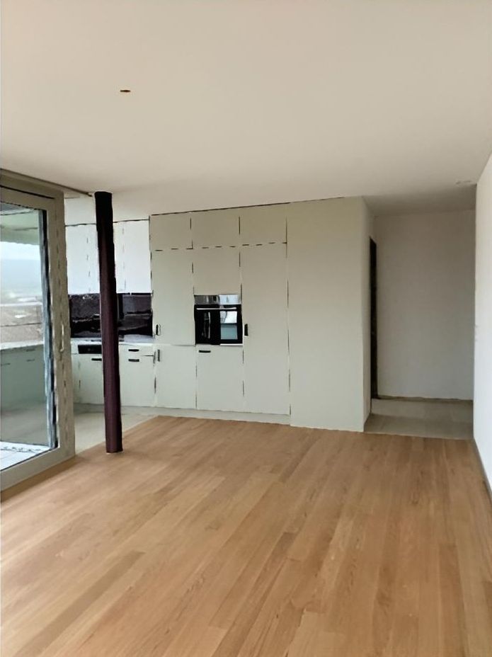 Ein heller, leerer Raum mit hellen Holzböden, einer modernen Küche mit weissen Schränken und einer dunklen Stützsäule.