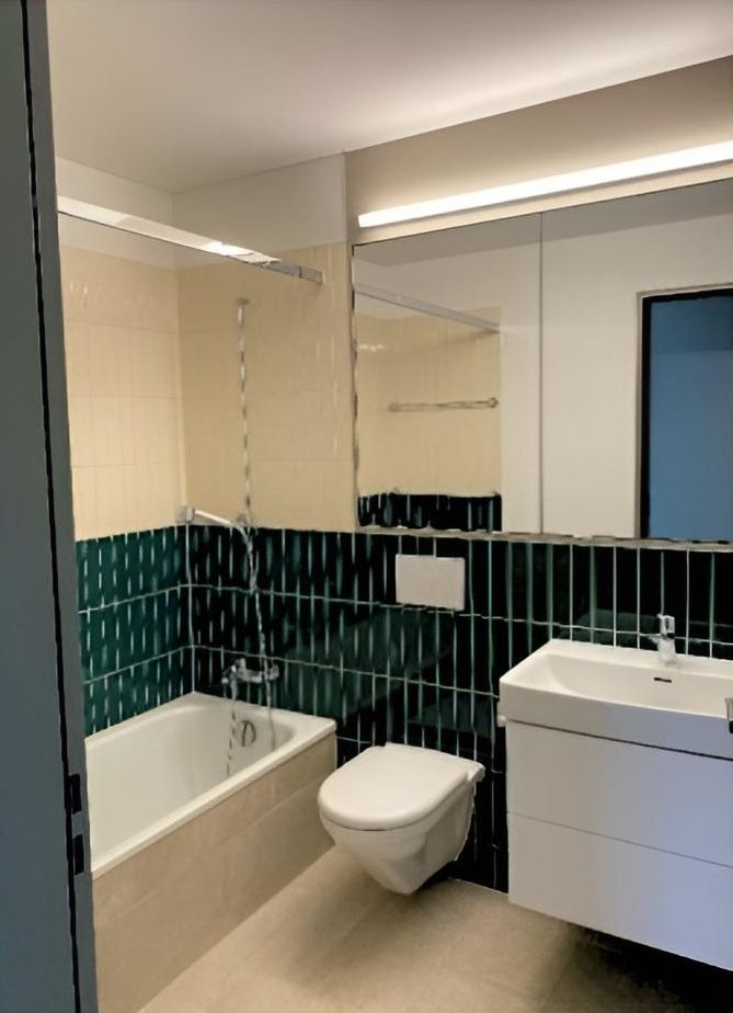 Modernes Badezimmer mit weisser Badewanne, wandhängendem WC, Waschtisch und grün gefliesten Wänden vor cremefarbenen Akzenten.