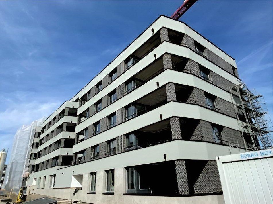 Ein mehrstöckiges Wohngebäude im Bau mit weissen horizontalen Streifen und grauer Backsteinfassade unter blauem Himmel.