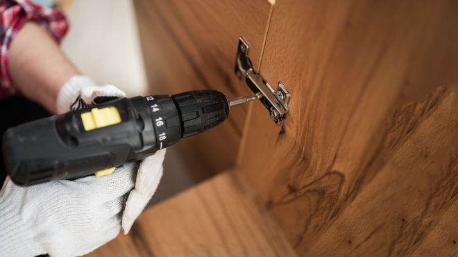 Eine Person mit Handschuhen montiert mit einer Bohrmaschine ein Scharnier an einem Holzschrank.