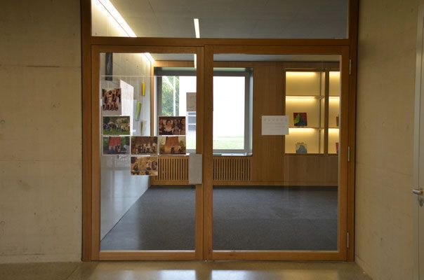 Doppelflügelige Holztüren führen in einen Raum. Links an der Wand hängen Fotografien, rechts stehen Vitrinen.