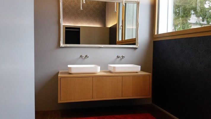 Badezimmer mit Waschtisch aus Holz, zwei weissen Waschbecken, grossem Spiegel und Fenster mit Tageslicht.