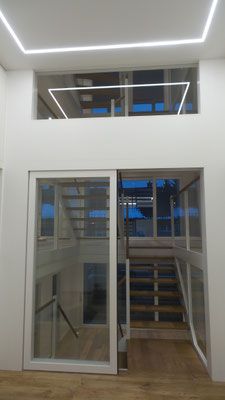 Innenansicht eines modernen Gebäudes, in dem hinter Glastüren und Fenstern ein Treppenhaus sichtbar ist. Darüber eine rechteckige Leuchte.