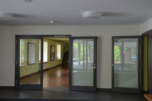 Flur mit drei Glastüren. Die offene Tür führt in einen Korridor mit Fenstern. Der Boden ist dunkel.