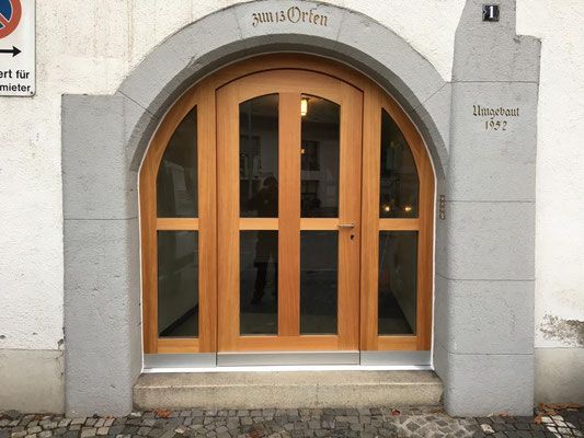 Hölzerner Bogeneingang mit Glaseinsätzen, eingelassen in eine Steinfassade. „WundOrle“ über der Tür.