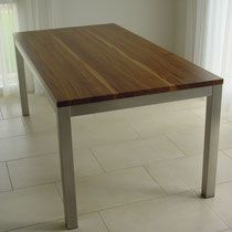 Ein rechteckiger Tisch mit Holzplatte und Metallbeinen steht auf einem gefliesten Boden, in der Nähe eines Fensters mit dünnen Vorhängen.