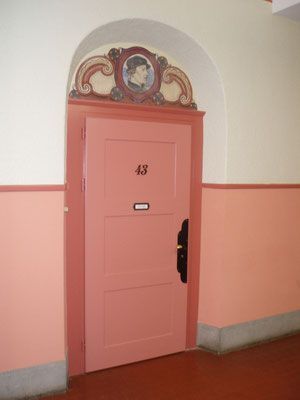 Eine rosafarbene Tür mit der Hausnummer 43 in einem pfirsichfarbenen Flur. Über der Tür hängt ein Porträt in einem Zierbogen.